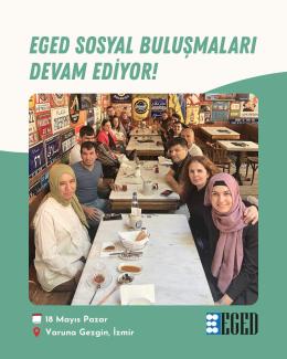 Bir kafede uzun bir masanın etrafında oturan yaklaşık on iki kişilik bir grup. Masada çay bardakları, tabaklar, zeytin ve ekmek gibi kahvaltılıklar var. İnsanlar birbirine yakın oturmuş, çoğu kameraya bakıyor ve gülümsüyor. Arka planda duvarlarda çeşitli tabelalar ve dekoratif objeler asılı. Fotoğrafın üst kısmında büyük harflerle “EGED SOSYAL BULUŞMALARI DEVAM EDİYOR!” yazıyor. Alt kısımda ise 18 Mayıs Pazar günü, Varuna Gezgin, İzmir’de buluşmanın gerçekleştiği belirtilmiş.