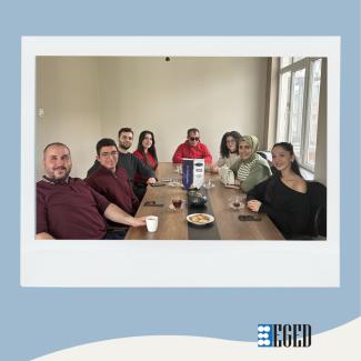 Bir toplantı masası etrafında oturan bir grup resmi  Ön planda soldan sağa: •Koyu bordo uzun kollu tişört giymiş, sağında bir beyaz fincan tutan sakallı erkek (gülümseyen) •Bordo kazaklı, gözlüklü genç erkek •Koyu sweatshirt giymiş sakallı genç erkek •Uzun koyu saçlı, kırmızı üstlü genç kadın  Masanın ortasında: •Gözlük takan kırmızı üstlü erkek (elleri masada) •Masada bir dikey duran beyaz-lacivert masa tabelası veya reklam kartı görünüyor.