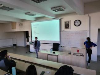 U düzenindeki toplantı masalarında oturmakta olan katılımcıların arkasından sahneyi görecek şekilde çekilmiş fotoğraf. Sahnede kır, kıvırcık saçlı tekerlekli sandalye kullanıcısı ellilerinde bir erkek yer almakta. Arkasında 1.70 - 1.80 metre yüksekliğinde bir tripod bulunmakta. Hemen sağındaki açık kahverengi örtülü panel masasında gür, kıvırcık saçlı, kırklarında gözlük kullanan kumral bir erkek oturmakta. Soldaki duvarda biri büyük ve biri küçük iki tablo görünmekte.