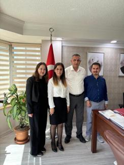 Fotoğrafta dört kişi bir ofis ortamında ayakta duruyor. Soldan sağa doğru: Seda, uzun saçlı, siyah ceket ve pantolon giymiş. Zeynep, uzun saçlı, beyaz gömlek ve siyah etek giymiş. Genel Müdür Cevdet Bey, Kısa saçlı, beyaz gömlek ve gri pantolon giymiş. Oğuzhan, kısa saçlı, mavi gömlek ve gri pantolon giymiş. Arka planda Türk bayrağı ve Atatürk'ün iki farklı portresi bulunuyor. Sol tarafta büyük bir saksıda yeşil yapraklı bir bitki var. Sağ tarafta ise bir masa üzerinde çeşitli evraklar ve dosyalar var.