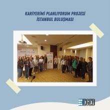 Konferans salonu, sunum yapılan bir etkinlik. Fotoğrafın ön planında bir mikrofon, bir not defteri ve bir kalem bulunan beyaz bir masa göze çarpıyor. Not defterinin üstünde "Zonat Hotel Edirne" yazısı var. Arka planda, bir kürsüde duran iki kişi izleyicilere hitap ediyor. Dinleyiciler ise yuvarlak bir masa etrafında oturmuşlar ve konuşmacıları dinliyorlar. Oda ferah, duvarlarda büyük pencereler ve floresan aydınlatmalar mevcut. Genel atmosfer toplantı veya eğitim etkinliği ortamında.