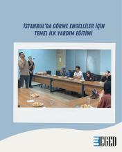Mavi arka plan üzerine yerleştirilmiş bir eğitim afişi. Üst kısımda koyu lacivert büyük harflerle şu metin yazıyor: “İSTANBUL’DA GÖRME ENGELLİLER İÇİN TEMEL İLK YARDIM EĞİTİMİ”.  Ortada beyaz çerçeveli bir fotoğraf var. Fotoğrafta dikdörtgen bir toplantı masasının etrafında oturan kişiler görülüyor. Masanın sol tarafında ayakta duran, kısa saçlı, açık renk kareli gömlekli bir erkek elinde mikrofonla konuşma yapıyor. Hemen yanında masanın üzerinde iki adet insan vücudu mankeni duruyor.
